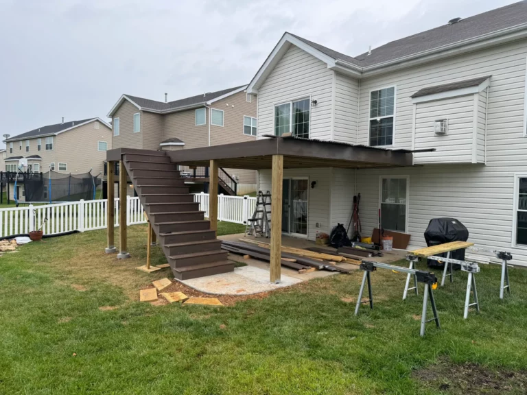 A partially constructed deck with wooden stairs leading to a house's backyard. The area shows tools and materials scattered around, with a grassy lawn and neighboring houses visible in the background.
