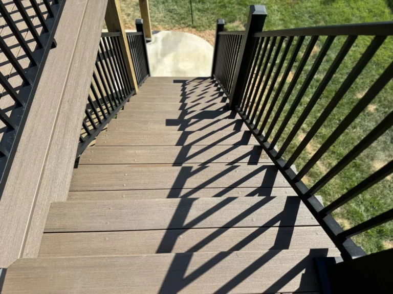 A view looking down a wooden staircase with black railings, casting shadows on the steps. The grass can be seen in the background.