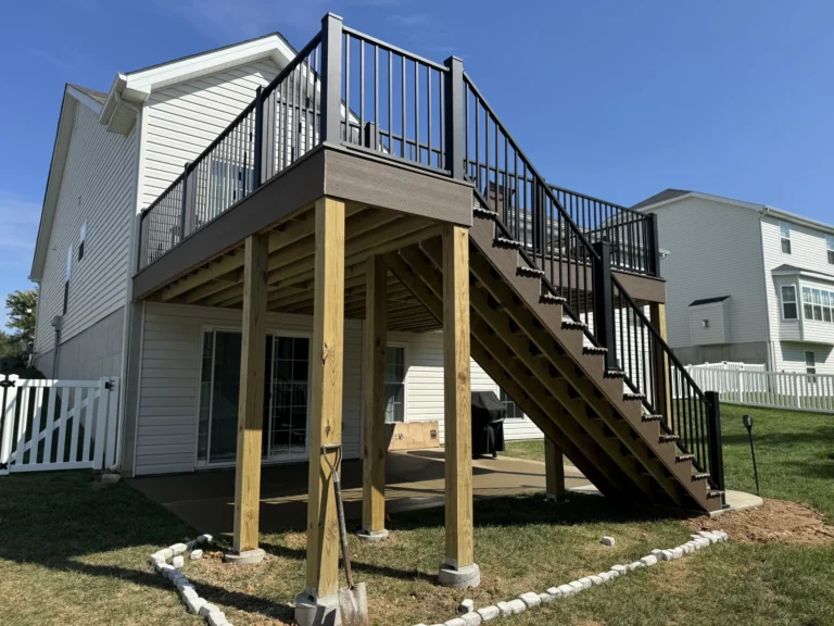 A two-story house with a large outdoor deck and stairs leading to the ground level. The deck has black railings and is partially shaded by the roof. The grassy yard has some decorative stones along the perimeter and a white fence in the background.
