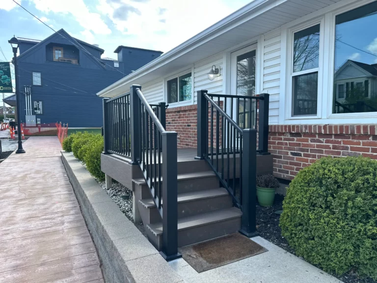 A side view of a house with a brick facade, featuring a set of wooden steps leading up to the entrance, surrounded by neatly trimmed bushes and a paved pathway.