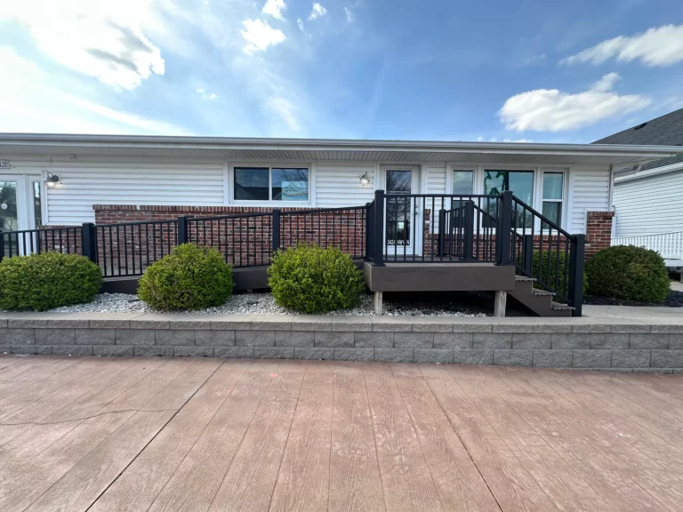A view of a ranch-style house with a brick façade, featuring a black railing staircase leading to the entrance. Lush green shrubs are planted at the base and a smooth concrete walkway is in the foreground under a partly cloudy sky.