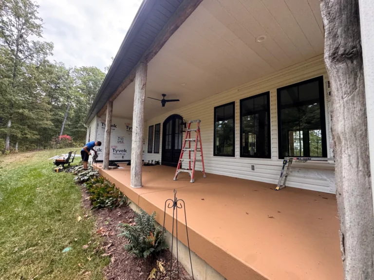 A person working on the porch of a house, surrounded by green grass and trees. The porch features large windows and wooden support beams, with a ladder and tools nearby.
