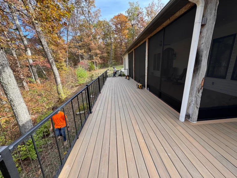 A spacious wooden deck with a black railing, surrounded by colorful autumn trees. A person in an orange shirt is walking nearby, while another person is visible further down the deck. The sky is bright and clear.
