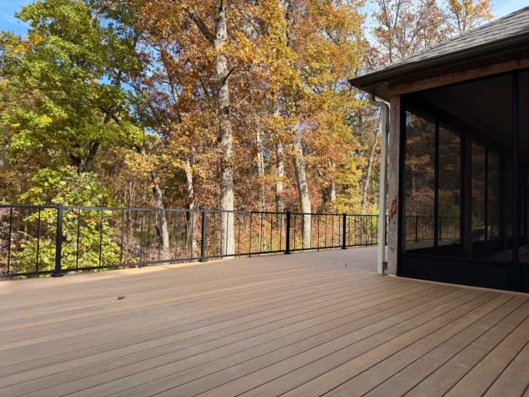 A spacious wooden deck surrounded by trees with autumn leaves, featuring a black railing and a sunlit view of the forest.