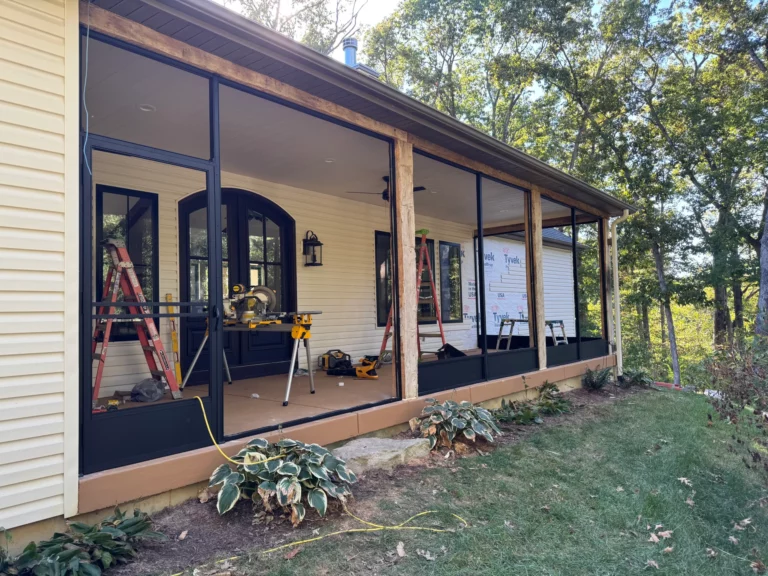 A partially completed screened-in porch featuring wooden framing and large windows. There are ladders and tools visible inside, along with greenery surrounding the base of the structure.