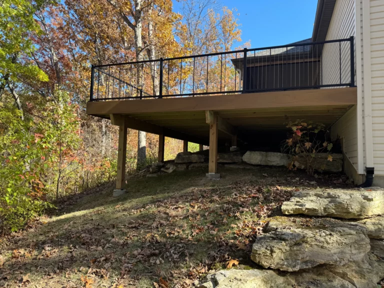 A wooden deck with a black railing extends from a house, positioned above a sloped yard covered with fallen leaves. In the background, colorful autumn trees are visible under a clear blue sky.